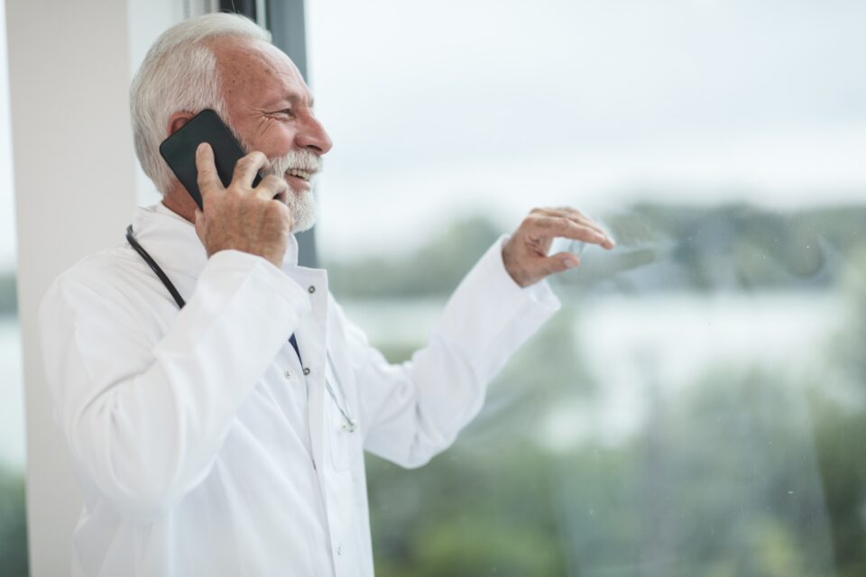 A physician in a white coat smiling on a phone call by a window.